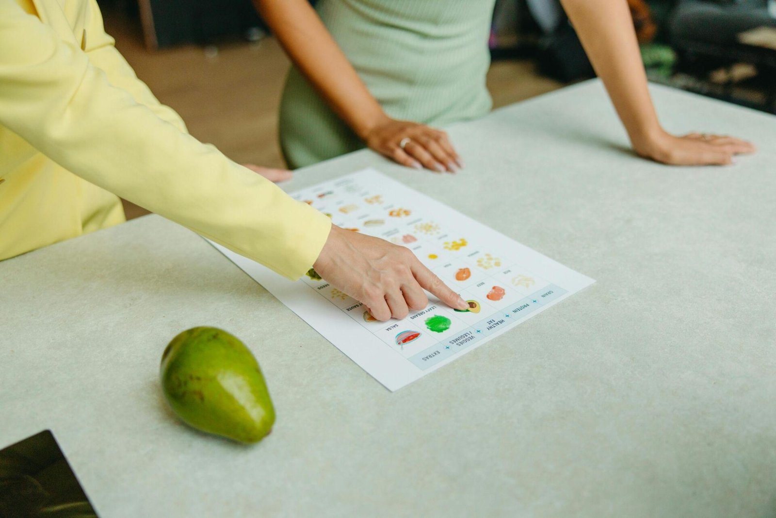 A medium close-up of a person in a white top and a bright pink shirt preparing a green smoothie in a kitchen. They are adding a red strawberry or piece of fruit into a blender filled with leafy greens. The countertop is covered with ingredients, including a bowl of fresh strawberries, a small bowl of cherries, a plate of spinach or other leafy greens, an orange/grapefruit, bottles of milk/liquid, and a brown glass bottle of supplements or vitamins. The kitchen has a modern, light aesthetic with blue cabinets.