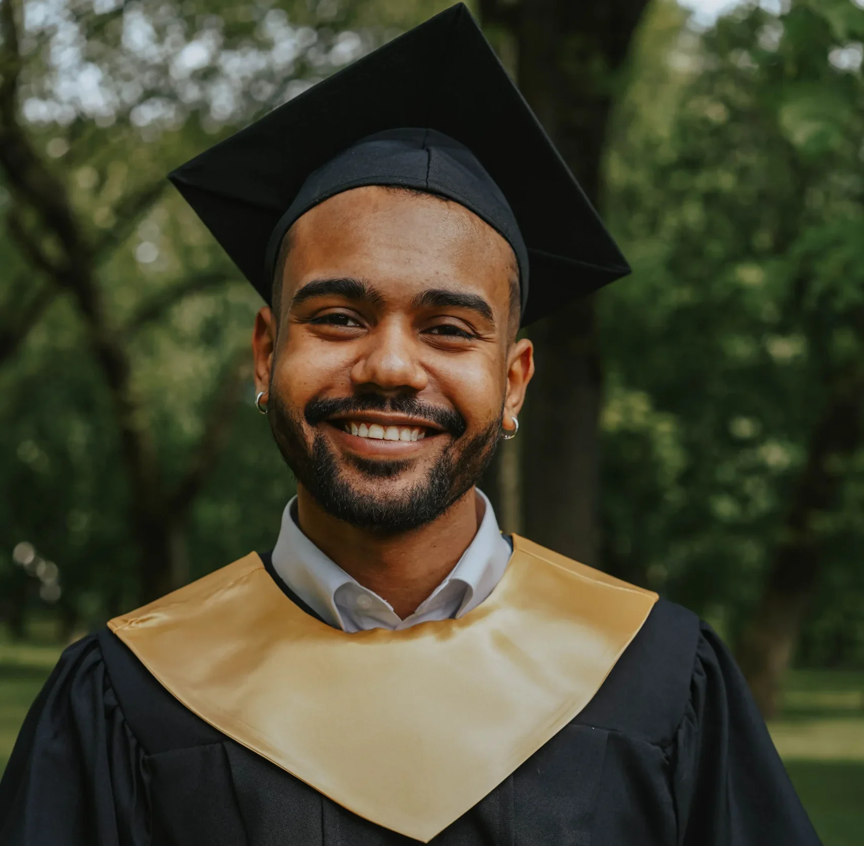 A smiling young man in a black graduation cap and gown with a gold stole posing outdoors.