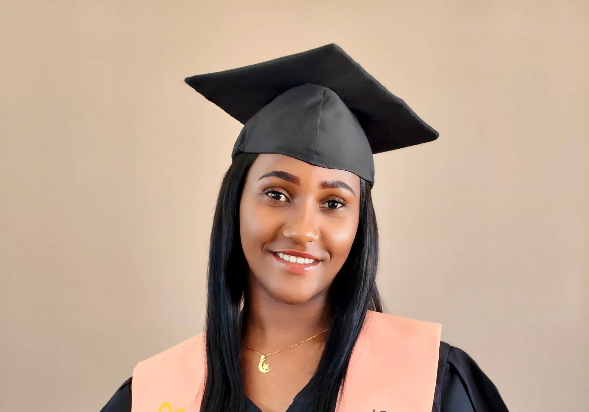 A smiling young woman in a black graduation cap and gown with a peach-colored stole.