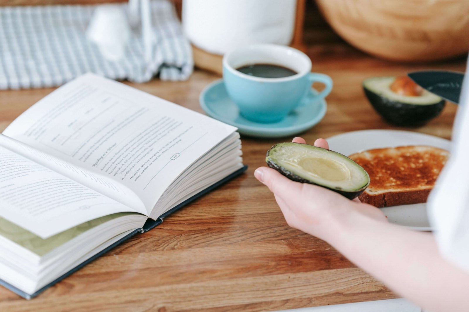 A person holding an avocado half over a piece of toast on a wooden table next to an open book and a blue cup of coffee.
