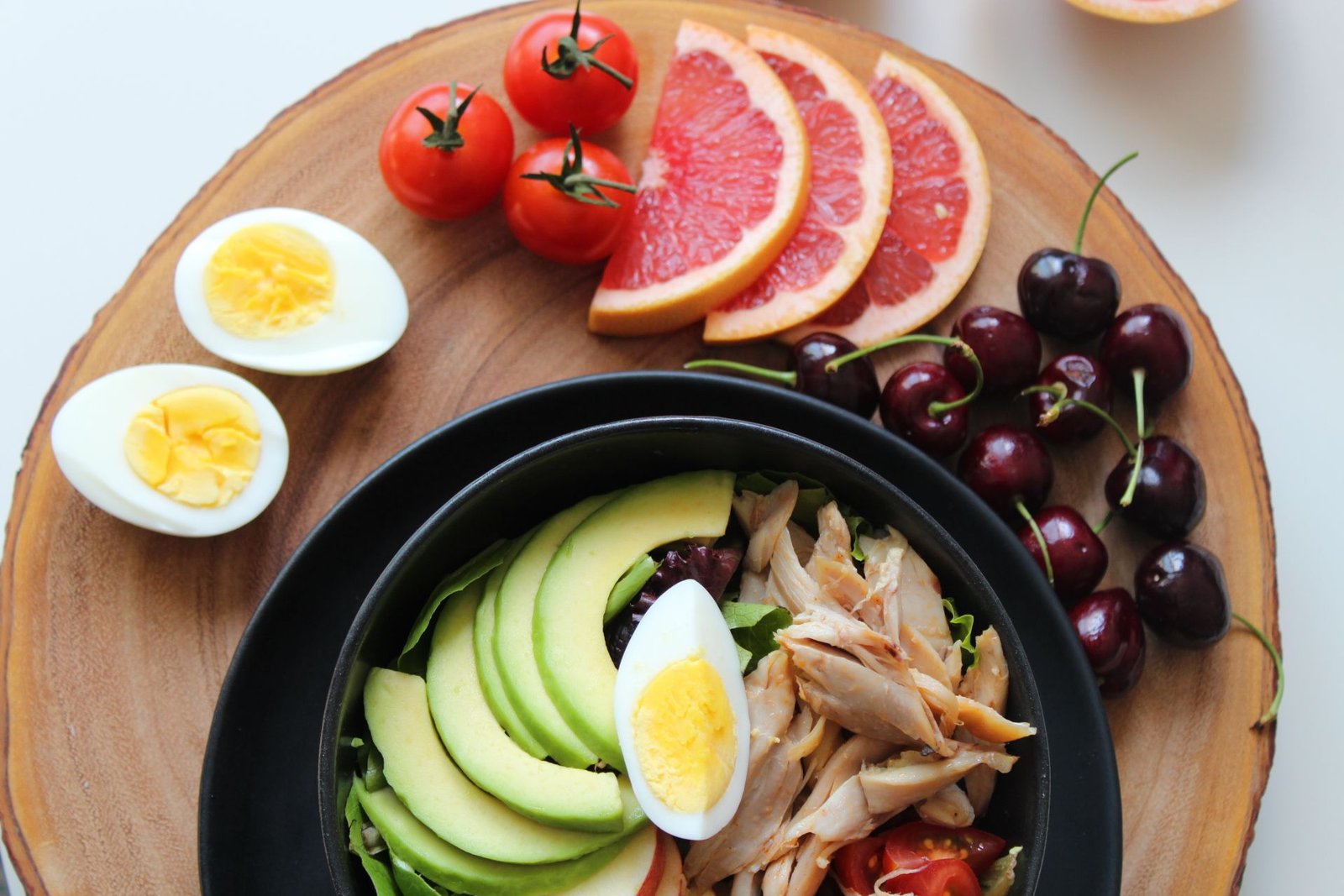 A healthy meal platter featuring a salad bowl topped with sliced avocado, chicken, and a hard-boiled egg, served on a wooden board with cherry tomatoes, grapefruit slices, cherries, and boiled egg halves