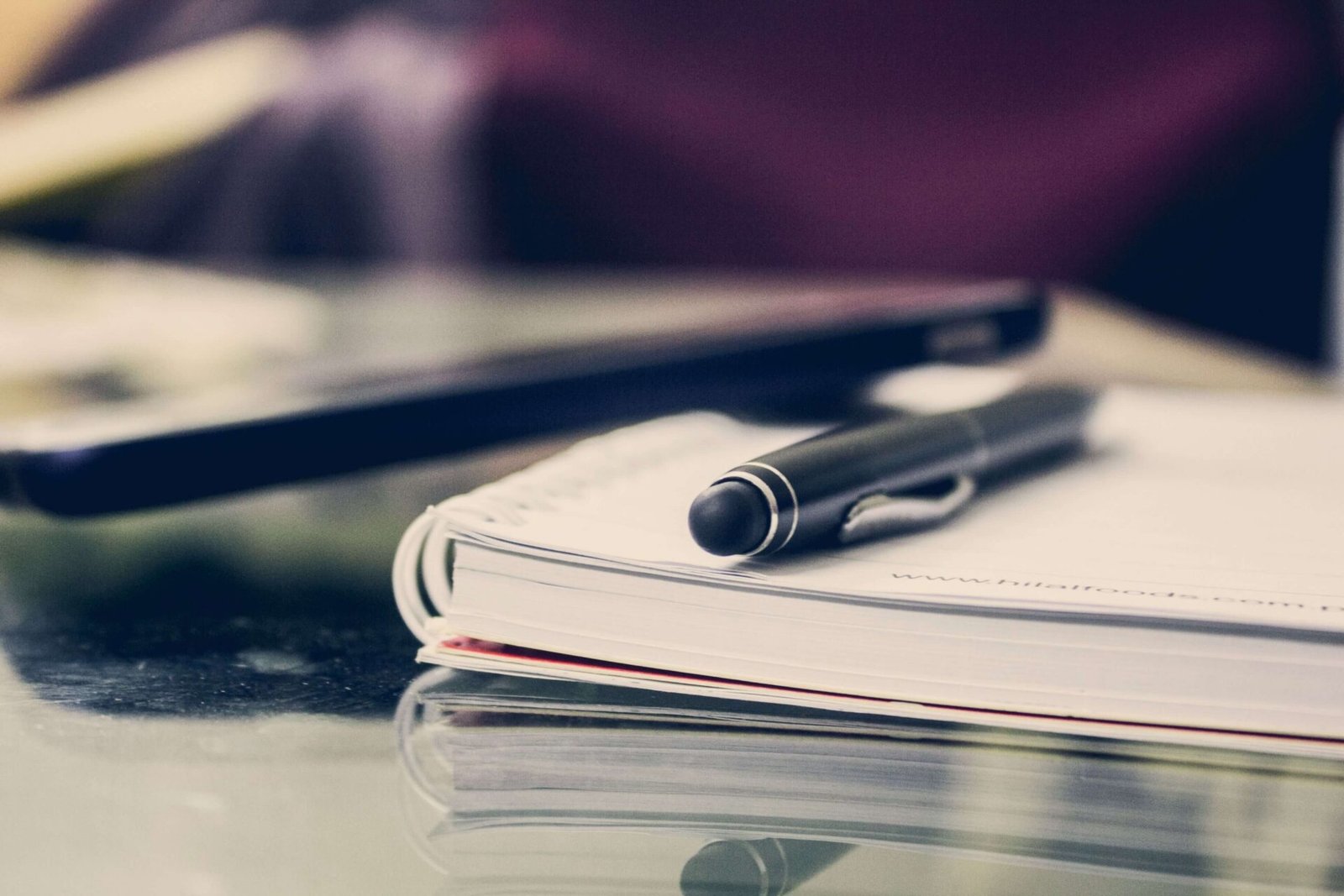 Black pen resting on top of an open notebook next to a smartphone on a reflective desk.