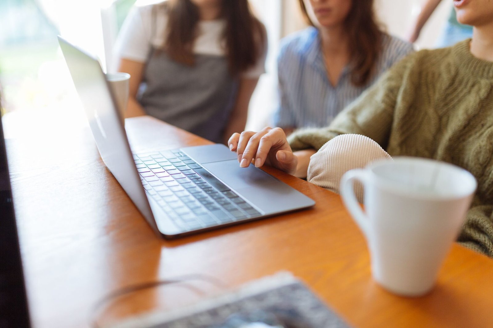 Close-up of a person typing on a laptop trackpad at a wooden counter next to a white coffee mug, with colleagues gathered in the background.
