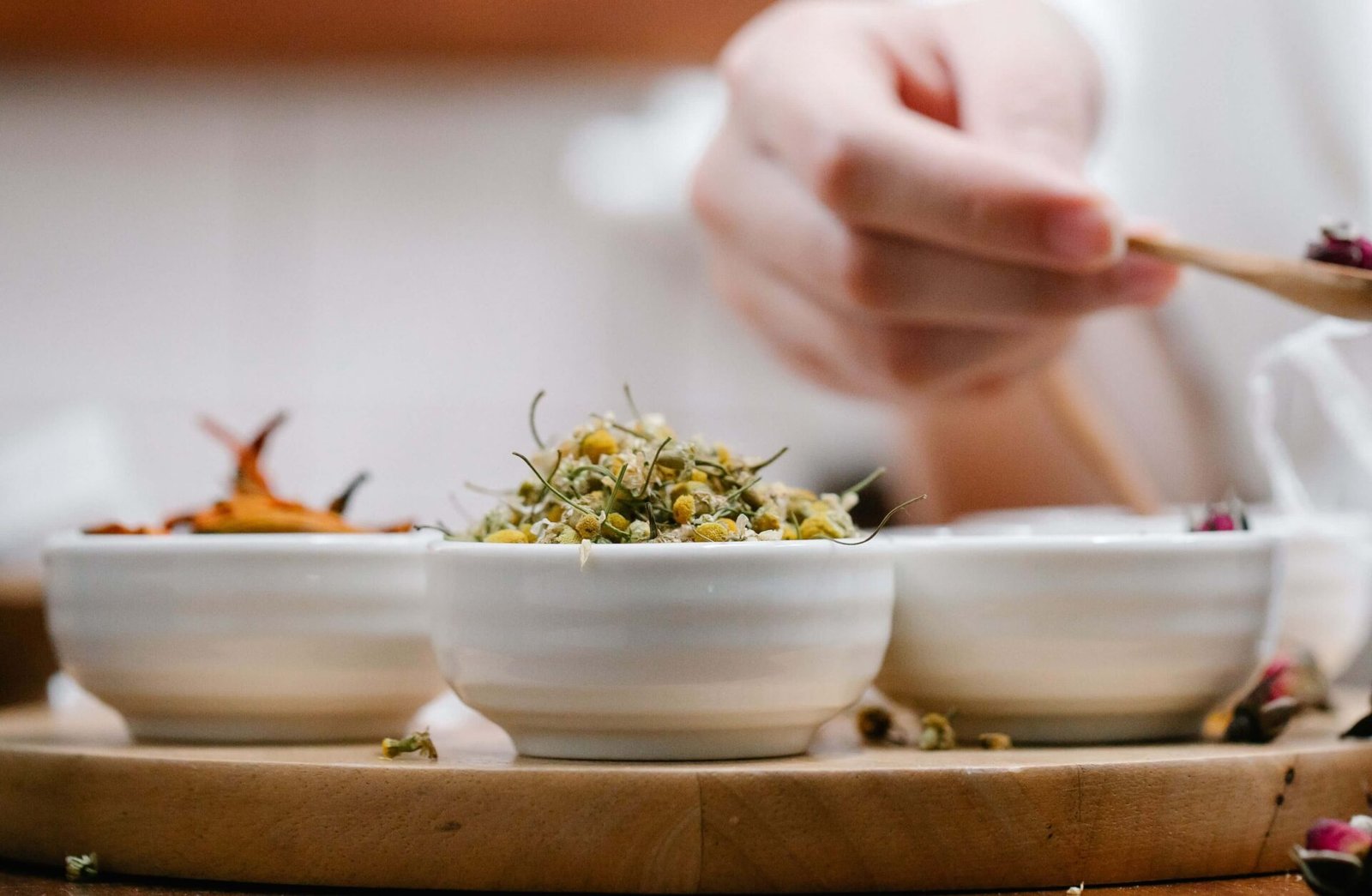 Close-up of small white bowls filled with dried chamomile and botanicals on a wooden board, with a hand holding a wooden spoon in the background