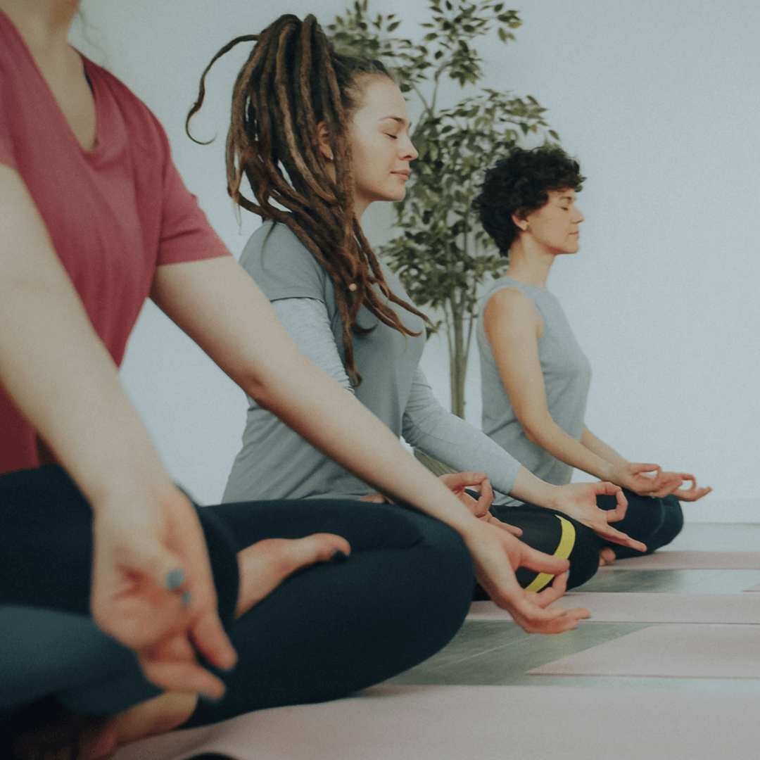 Group of people sitting cross-legged on yoga mats in a quiet studio, practicing meditation with eyes closed and hands in a mudra position.