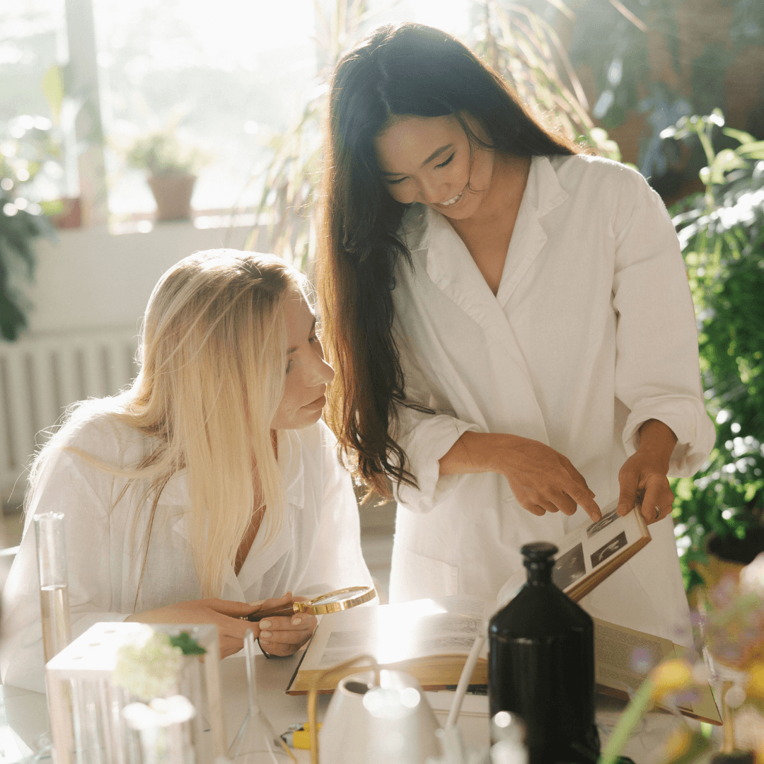 Two female researchers in white lab coats reviewing notes and discussing data together in a sunlit, plant-filled laboratory.
