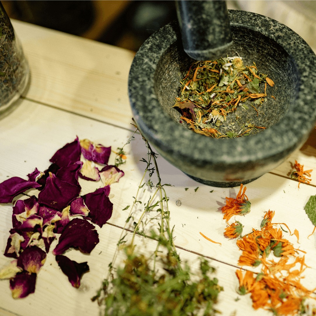 High-angle shot of a granite mortar and pestle grinding herbs next to scattered rose petals and orange marigold flowers on a wooden table.
