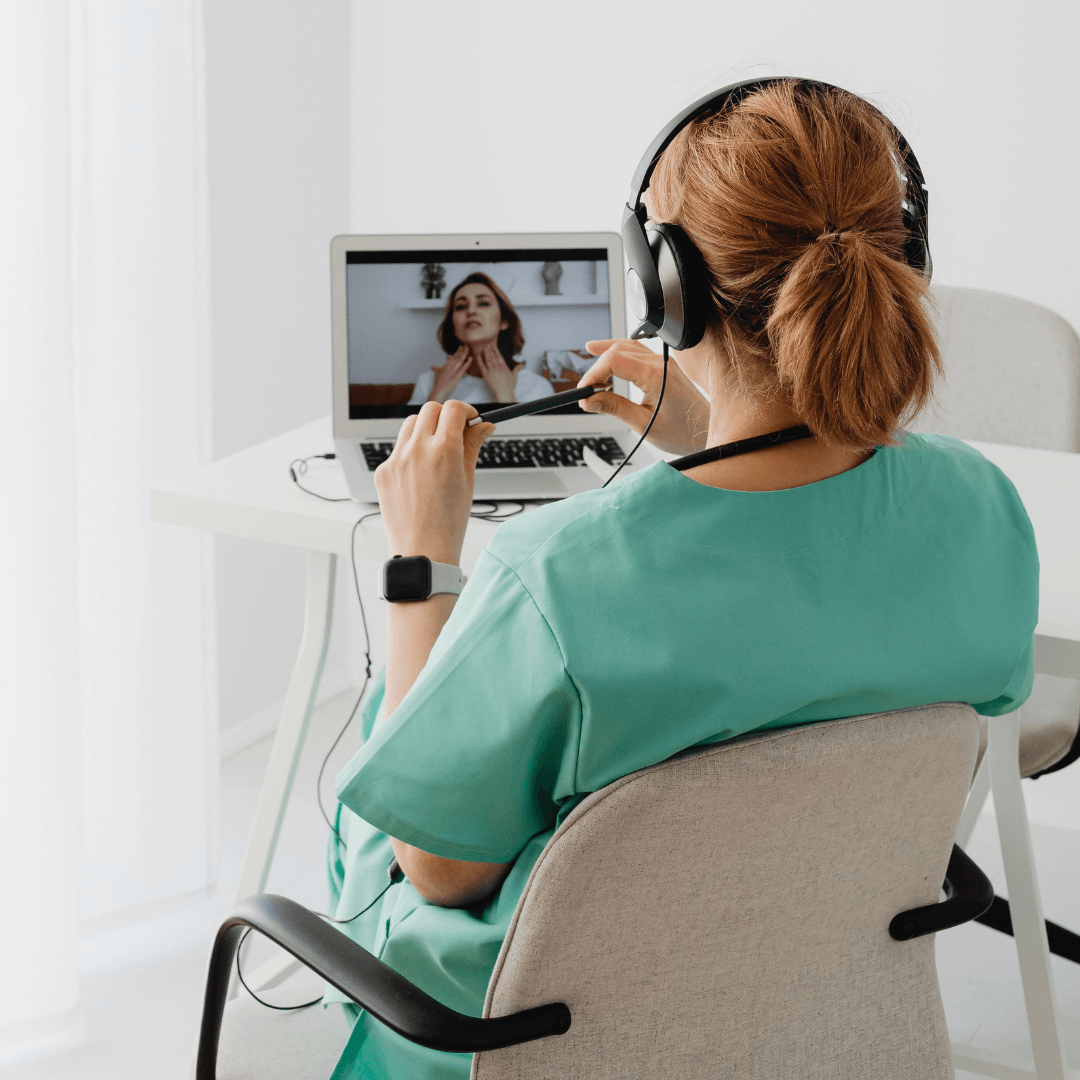 Rear view of a healthcare professional in green scrubs and a headset conducting a telehealth consultation on a laptop with a patient