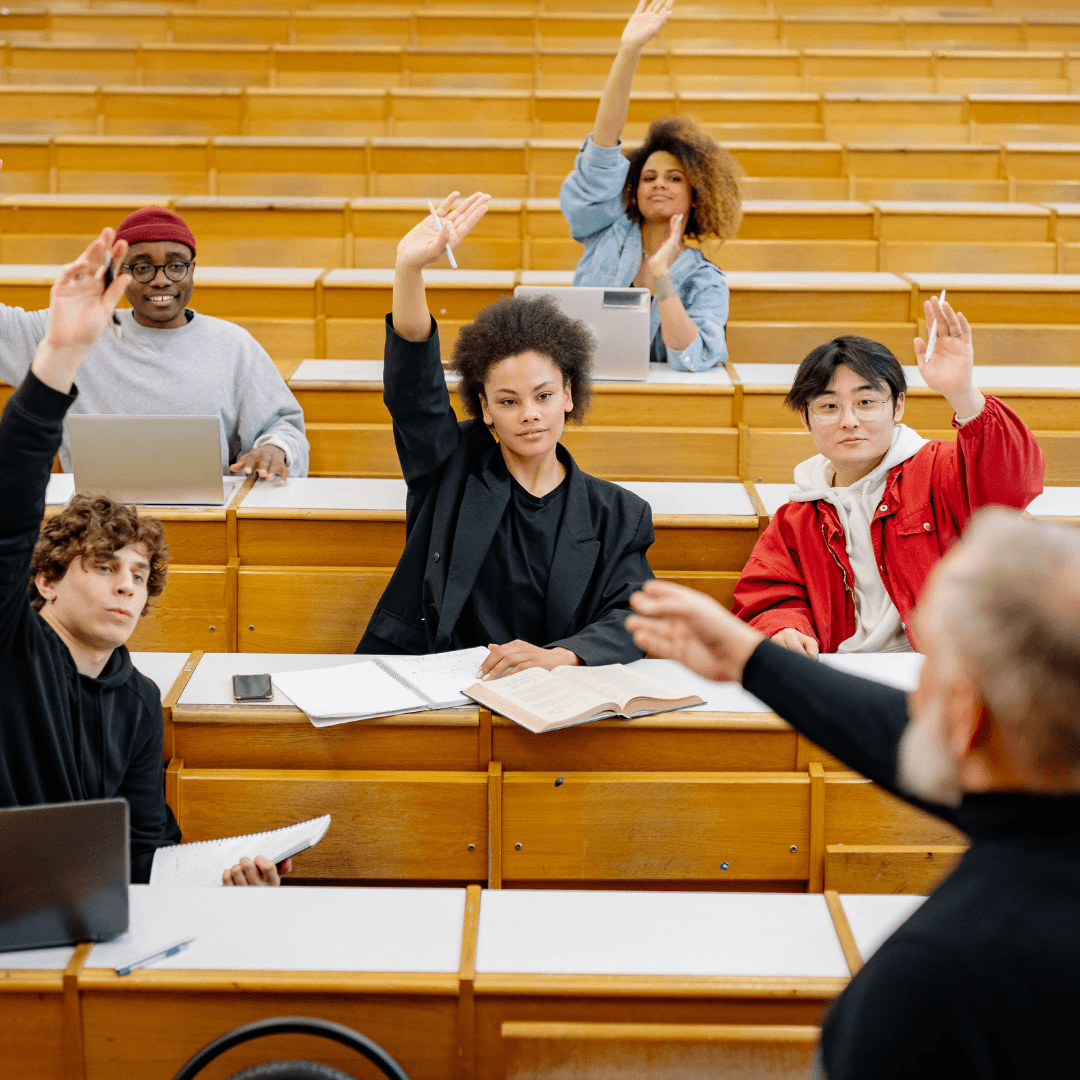 Diverse group of university students raising their hands in a large tiered lecture hall to answer a professor's question